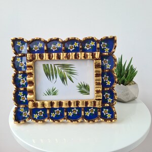 Pu&ograve; includere: Una cornice per foto blu e oro con un bordo a festoni e motivi floreali. La cornice &egrave; decorata con fiori bianchi e foglie verdi.