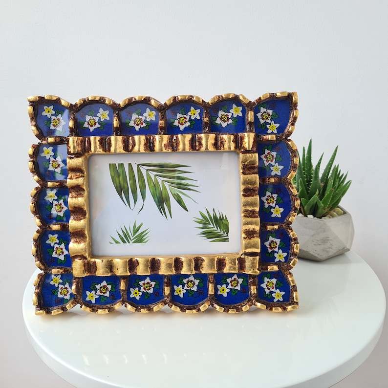 Pu&ograve; includere: Una cornice per foto blu e oro con un bordo a festoni e motivi floreali. La cornice &egrave; decorata con fiori bianchi e foglie verdi.