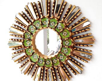 May include: A round sunburst mirror with a gold frame and green and white tile-like design. The mirror has a circular reflection.