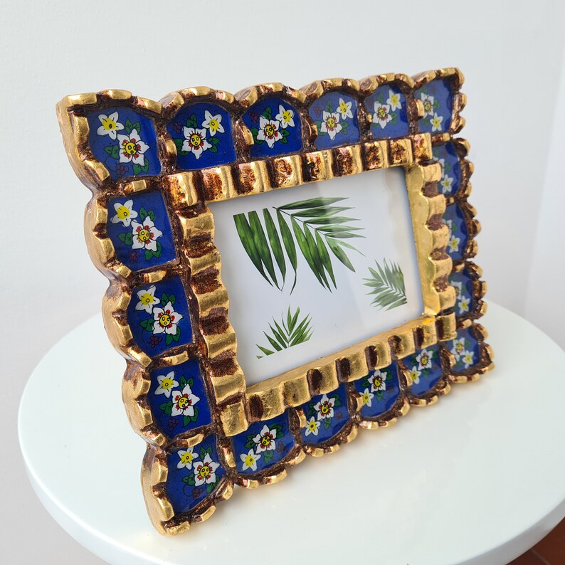 Pu&ograve; includere: Una cornice dorata con un design floreale blu e bianco. La cornice ha un bordo a festoni ed &egrave; decorata con piccoli fiori bianchi. La cornice &egrave; vuota e pronta per contenere una foto.