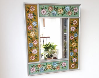 May include: A silver framed mirror with a floral design. The frame is decorated with painted flowers in shades of pink, blue, yellow, and green on a gold background.