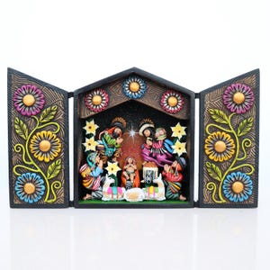 May include: A colorful, hand-painted nativity scene in a triptych design. The wooden frame features floral patterns and opens to reveal the holy family, angels, and animals. The scene is set against a starry background.