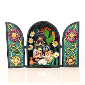 May include: A colorful, hand-painted nativity scene with a black backdrop and floral accents. The scene features figures of Mary, Joseph, and baby Jesus, with an angel and other figures. The side panels have large pink and yellow flowers.