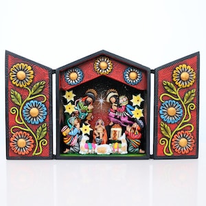 May include: A colorful, three-panel nativity scene. The central panel depicts the holy family, surrounded by figures and animals. The panels are framed in black and decorated with red, yellow, blue, and green floral designs. The scene is set against a white background.