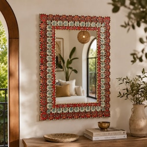 Puede incluir: Un espejo de pared rectangular con un marco decorativo festoneado con paneles alternos rojos y blancos. El marco está adornado con intrincados patrones florales en tonos azules, verdes y amarillos. El espejo refleja el interior de una habitación.