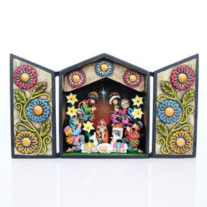 May include: A three-panel, hand-painted nativity scene. The central panel depicts the holy family and onlookers, while the side panels feature floral designs with pink, blue, and yellow flowers. The overall color palette includes earth tones and vibrant accents.