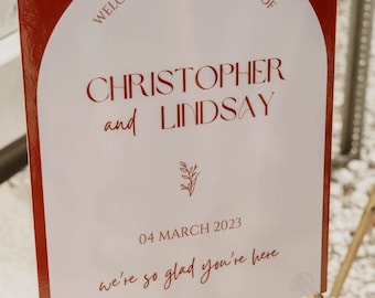 May include: A white welcome sign with red text and a red border. The sign reads "Welcome to the wedding of Christopher and Lindsay" with the date "04 March 2023" and the phrase "we're so glad you're here".