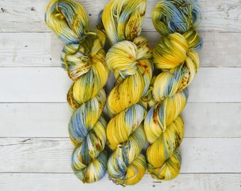 Op de afbeelding: Drie bollen garen in tinten van blauw, geel en groen. Het garen is handgeverfd en heeft een gespikkeld uiterlijk.