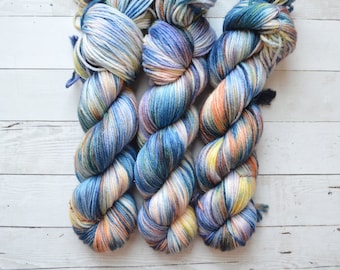 Op de afbeelding: Drie bollen garen in een blauwe, witte en oranje kleurstelling. Het garen is handgeverfd en heeft een gemêleerd patroon.