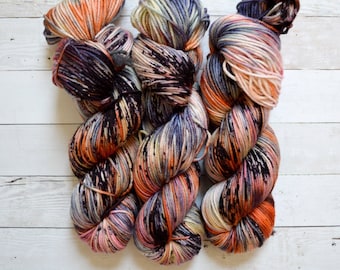 May include: Three skeins of hand-dyed yarn in a variety of colors. The yarn features shades of orange, purple, pink, and cream, with darker speckles throughout. The yarn is arranged on a white wooden surface.