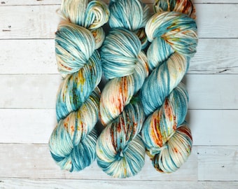 Op de afbeelding: Drie bollen garen in een blauwe, witte en oranje kleurstelling. Het garen is handgeverfd en heeft een gespikkeld uiterlijk.