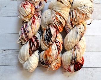 May include: Three skeins of hand-dyed yarn in a cream base with vibrant speckles of red, orange, black, and purple. The yarn is twisted into thick, soft-looking bundles, perfect for knitting or crocheting projects.