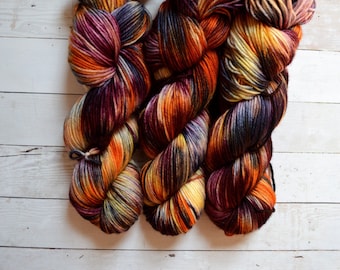 May include: Three skeins of yarn in a variegated colorway with shades of orange, yellow, brown, and purple. The yarn is arranged in a loose braid.