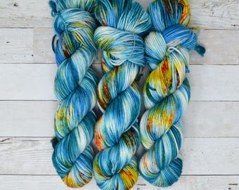 Op de afbeelding: Drie bollen garen in een blauwe, witte en gele kleurstelling. Het garen is handgeverfd en heeft een gemêleerd patroon.