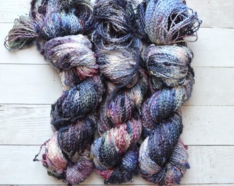 Könnte beinhalten: Drei Knäuel Garn in den Farben Lila, Blau und Schwarz mit weißen und silbernen Akzenten. Das Garn hat eine flauschige Textur und ist mit kleinen Perlen verziert.