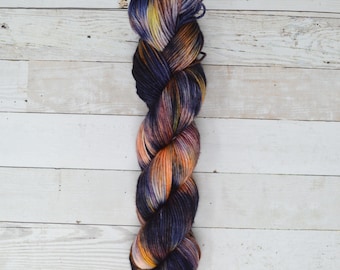 May include: A skein of yarn in a deep purple, orange, and yellow colorway. The yarn is twisted into a braid and is ready to be used for knitting or crocheting.