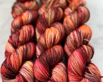 May include: A collection of vibrant yarn skeins in shades of red, orange, and brown. The yarn is tightly wound, showcasing a gradient of colors. The skeins are arranged closely together, highlighting the texture and color variations.