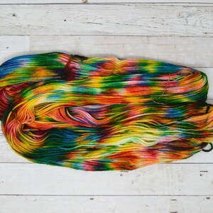 Puede incluir: Un ovillo de hilo de colores con tonos de verde, amarillo, naranja, rojo y azul. El hilo est&aacute; dispuesto en una pila suelta sobre una superficie de madera blanca.