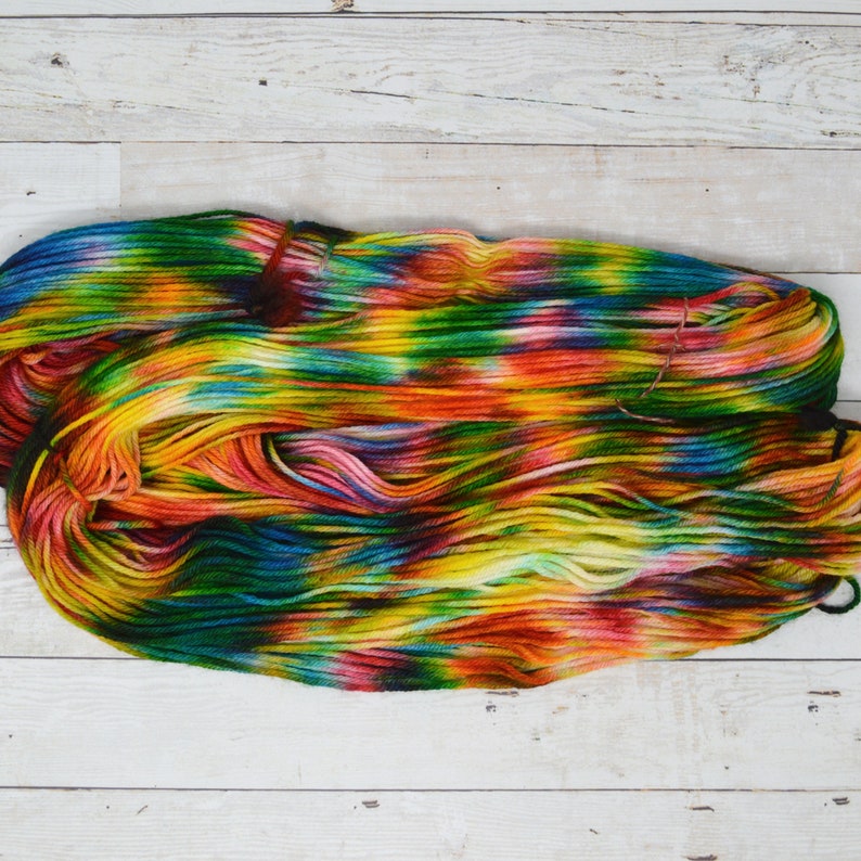 Puede incluir: Un ovillo de hilo de colores con tonos de verde, amarillo, naranja, rojo y azul. El hilo est&aacute; dispuesto en una pila suelta sobre una superficie de madera blanca.