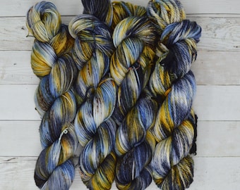 Op de afbeelding: Een verzameling van zes bollen garen in tinten blauw, geel en zwart. Het garen is handgeverfd en heeft een gespikkeld uiterlijk.