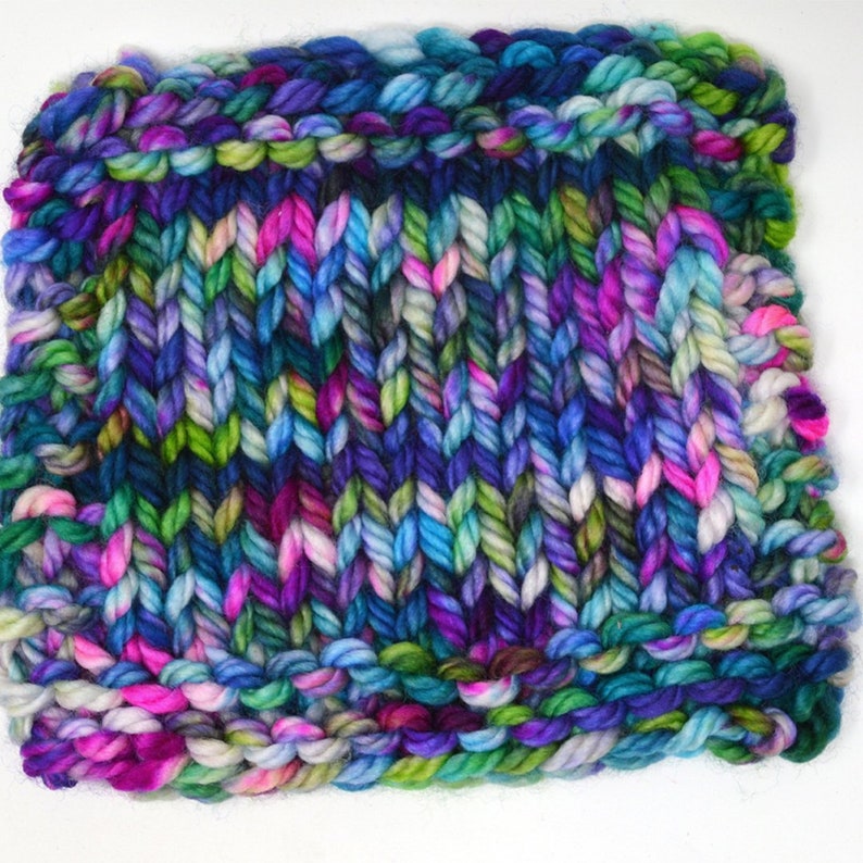 K&ouml;nnte beinhalten: Eine Nahaufnahme eines gestrickten Stoffmusters in verschiedenen Farben, darunter Blau, Gr&uuml;n, Pink und Lila. Das Garn ist dick und strukturiert und erzeugt einen grob gestrickten Look.