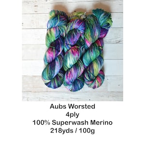 K&ouml;nnte beinhalten: Drei Kn&auml;uel buntes Garn in den Farben T&uuml;rkis, Gr&uuml;n, Lila, Rosa und Gelb. Das Garn ist mit "Aubs Worsted 4ply 100% Superwash Merino 218yds / 100g" beschriftet.