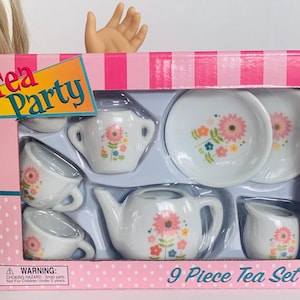 Puede incluir: Un juego de té de juguete de 9 piezas en una caja rosa. El juego incluye una tetera, una jarra de crema, un azucarero, dos platillos y cuatro tazas de té. Cada pieza blanca tiene un diseño floral. La caja dice "Tea Party" y "9 Piece Tea Set".