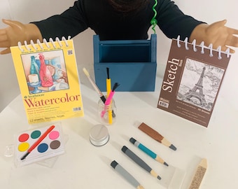 May include: A set of art supplies for kids, including a watercolor pad, a sketch pad, paintbrushes, a jar of water, a palette of watercolors, and pencils. The sketch pad features a drawing of the Eiffel Tower and the text "Sketch Book".