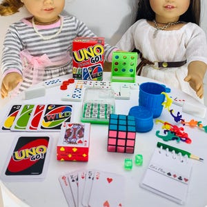 May include: A collection of children's games and toys including a red and blue UNO Go! card game, a green and white Connect 4 game, a clear plastic container with white letter tiles, a red and black Rubik's Cube, a blue plastic cup, a set of dominoes, a set of playing cards, and a set of colorful plastic toys.