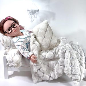 May include: A doll wearing glasses and pajamas, lying in a white bed with a fluffy blanket and pillow. The doll has brown hair with pink highlights. The bed has a white frame. A small chandelier hangs above the bed.