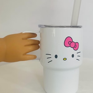 Puede incluir: Vaso blanco con diseño de Hello Kitty, que incluye un lazo rosa y rasgos faciales. Tiene un asa, una tapa transparente y una pajita transparente. El vaso está sostenido por una mano.