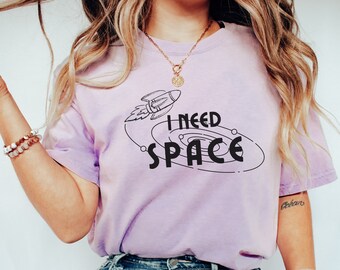 May include: A woman wearing a light purple t-shirt with a black graphic of a rocket ship and the text "I NEED SPACE".