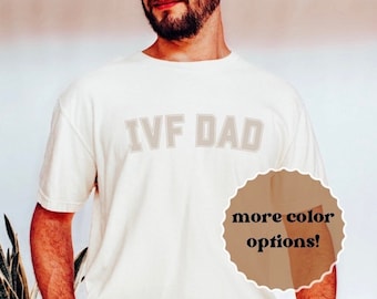 May include: A white t-shirt with the text "IVF DAD" printed in a light gray color. The shirt is being worn by a person with a beard. The text "more color options!" is written in a circle on the right side of the image.