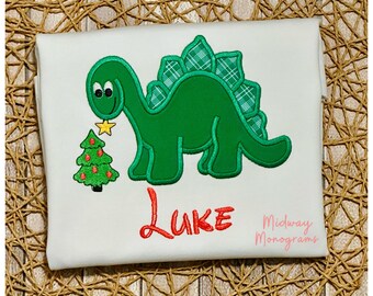 May include: A white t-shirt with a green dinosaur holding a small Christmas tree. The dinosaur has a plaid pattern on its back. The name "Luke" is embroidered in red below the dinosaur. The text "Midway Monograms" is in pink below the name.