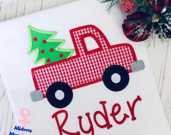 May include: White t-shirt featuring a red and white gingham truck carrying a green Christmas tree with red ornaments. The name "Ryder" is embroidered in red script. Black wheels and gray accents complete the design. A festive holiday theme.