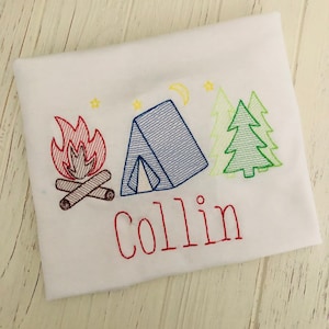 May include: A white t-shirt with an embroidered design of a campfire, tent, and pine trees. The name "Collin" is embroidered below the design in red thread.