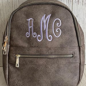 May include: A brown faux leather backpack with a front zipper pocket and a purple monogram "AMC" embroidered on the front.