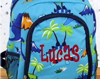 May include: Blue dinosaur backpack with a green, orange, and white pattern. The backpack has a zipper closure and a mesh pocket on the side. The name "Lucas" is embroidered in red on the front of the backpack.