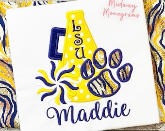 May include: A white t-shirt featuring an embroidered yellow megaphone with white polka dots, the letters LSU, a purple and yellow pom-pom, and a purple paw print with tiger stripes. The name Maddie is embroidered in purple script below the design.