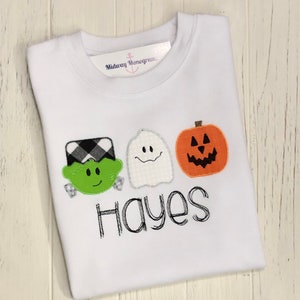May include: White long-sleeved shirt with a green Frankenstein, white ghost, and orange pumpkin appliqu&eacute;. The shirt has the name "Hayes" embroidered in black.