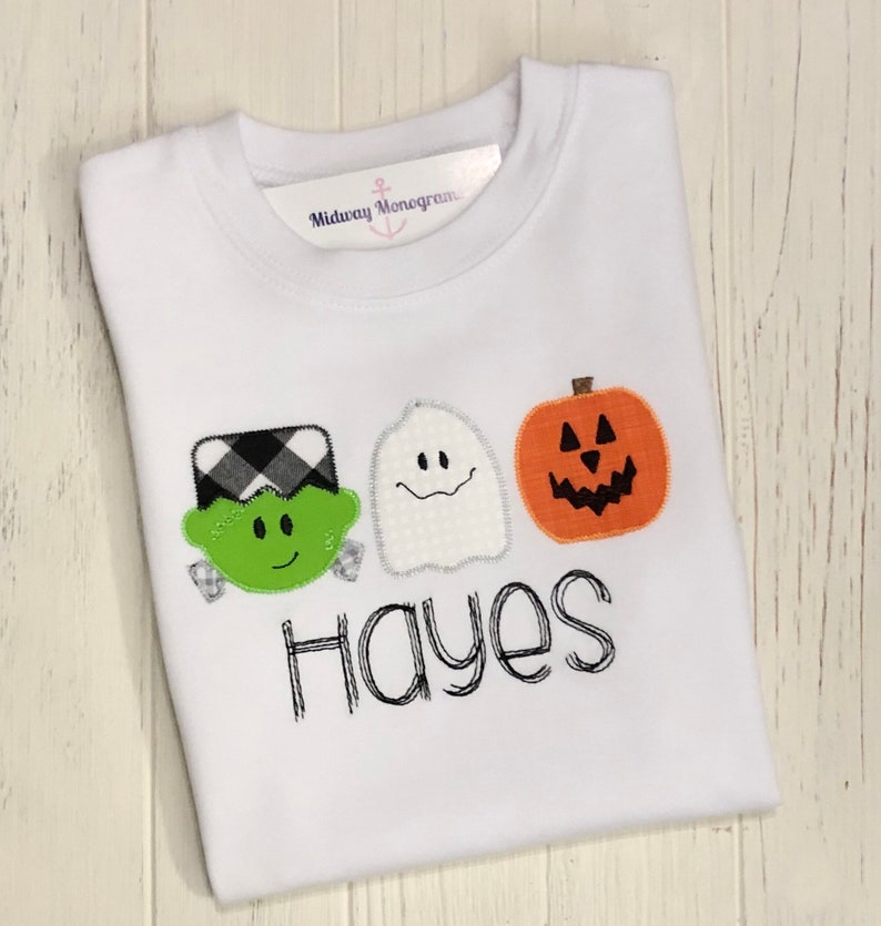 May include: White long-sleeved shirt with a green Frankenstein, white ghost, and orange pumpkin appliqu&eacute;. The shirt has the name "Hayes" embroidered in black.