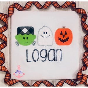 May include: White t-shirt featuring a green Frankenstein, a white ghost, and an orange pumpkin. The name "Logan" is embroidered beneath the characters.