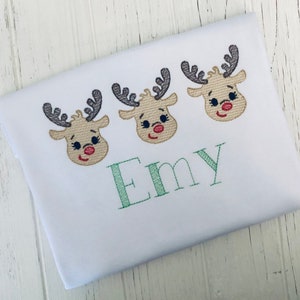 May include: White t-shirt with three embroidered reindeer faces and the name "Emy" in green thread.