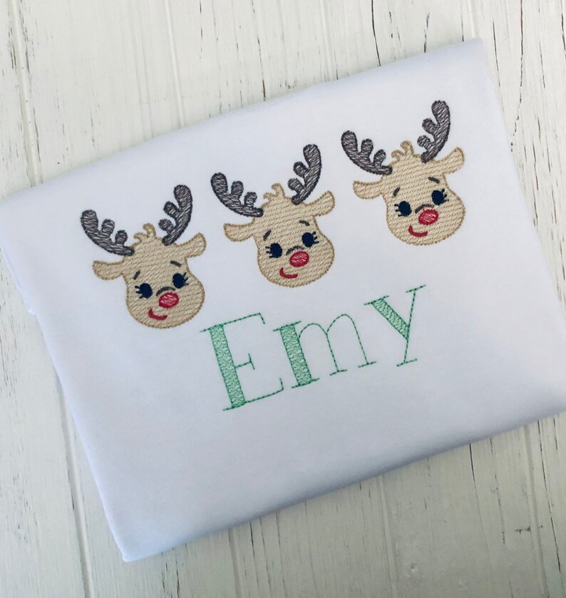 May include: White t-shirt with three embroidered reindeer faces and the name "Emy" in green thread.