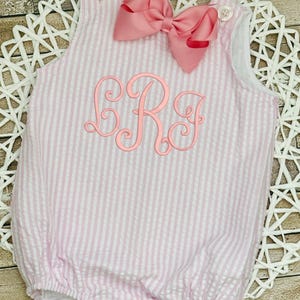 May include: Pink and white striped baby romper with a large pink bow and embroidered initials. The romper has button closures on the shoulders and bottom. The fabric has a seersucker texture.