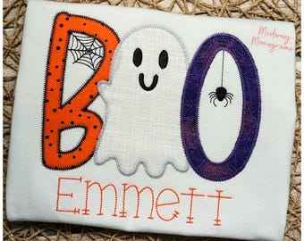 May include: White t-shirt with the word "BOO" in orange and purple, featuring a ghost and spider design. The name "Emmett" is embroidered in orange below the word "BOO". A Halloween-themed shirt.