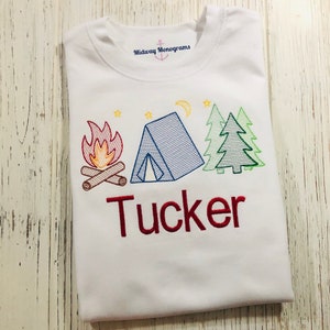 May include: White t-shirt with an embroidered design of a campfire, tent, and pine trees. The name "Tucker" is embroidered in red below the design. The text "Midway Monograms" is visible at the top of the shirt.