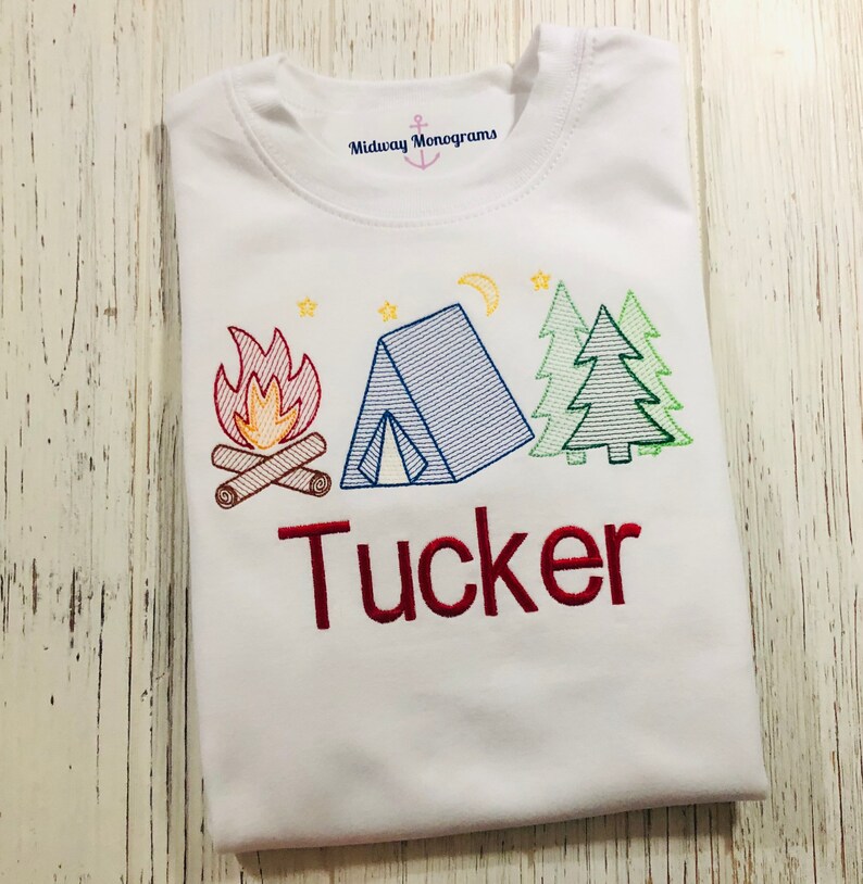 May include: White t-shirt with an embroidered design of a campfire, tent, and pine trees. The name "Tucker" is embroidered in red below the design. The text "Midway Monograms" is visible at the top of the shirt.
