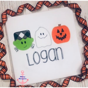 May include: White t-shirt featuring a green Frankenstein, a white ghost, and an orange pumpkin. The name "Logan" is stitched in black below the characters.
