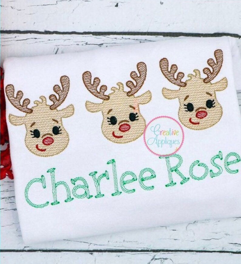 May include: White embroidered shirt with three red-nosed reindeer faces and the name "Charlee Rose" stitched in green thread.